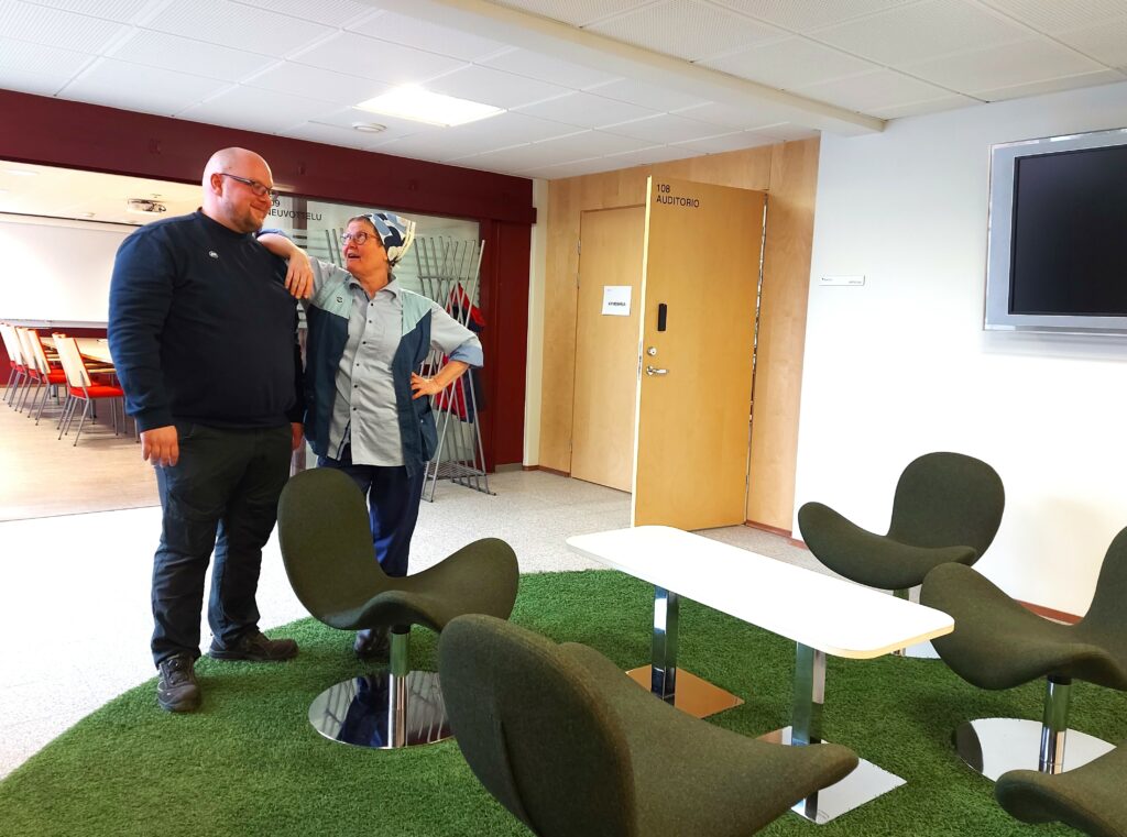 Nainen ja mies seisovat vierekkäin aulatilassa ja katsovat toisiaan.